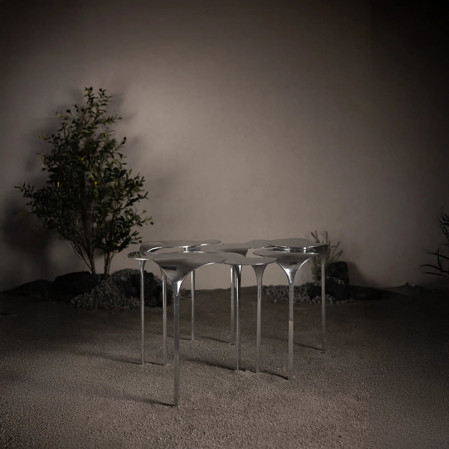 A sculptural coffee table made of aluminum with a modular design, displayed in a minimalist setting with a plant and a wall in the background.