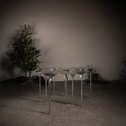 A sculptural coffee table made of aluminum with a modular design, displayed in a minimalist setting with a plant and a wall in the background.